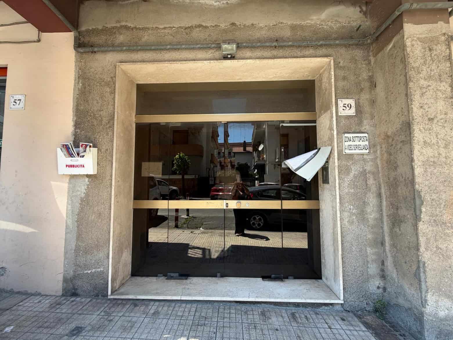 Ingresso di un edificio commerciale con porta in vetro e struttura in cemento, situato in una zona urbana con veicoli parcheggiati e edifici circostanti.