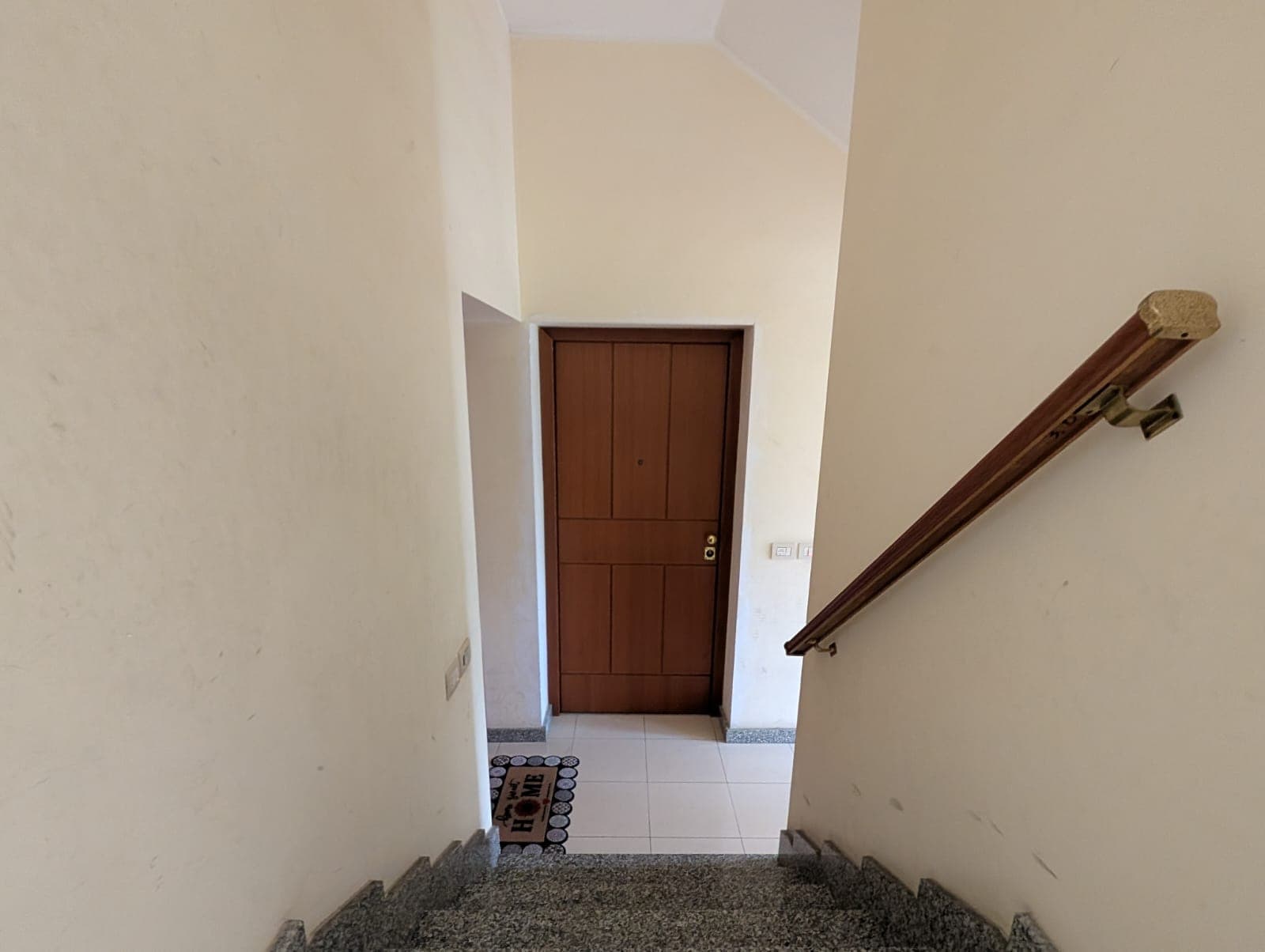 Aggiunta di un ingresso con porta in legno e scala interna in un'abitazione, vista dall'alto con pareti chiare e ringhiera in legno.