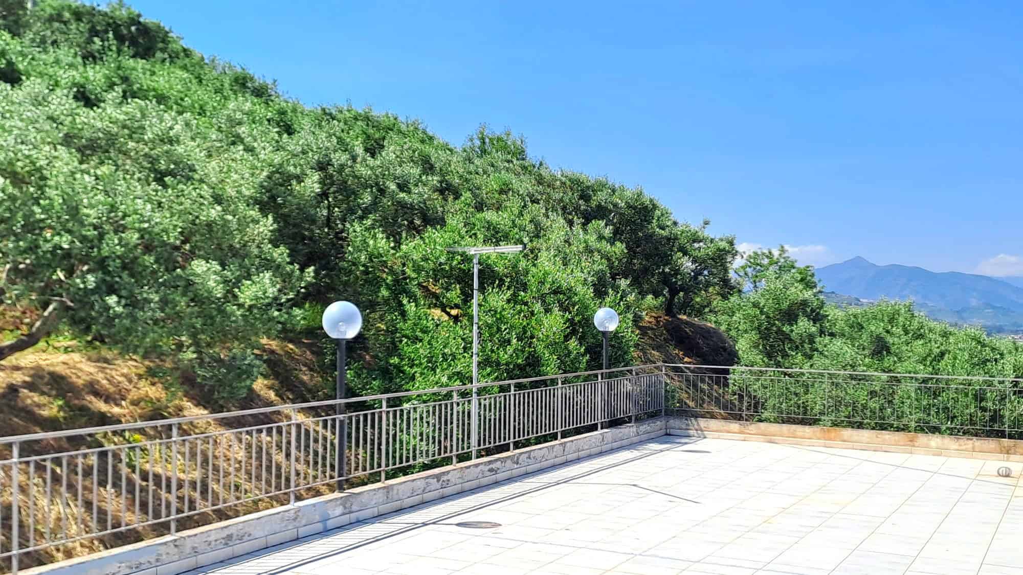 Vista panoramica di un terrazzo con ringhiera in metallo, circondato da vegetazione verde e colline sullo sfondo, sotto un cielo azzurro.
