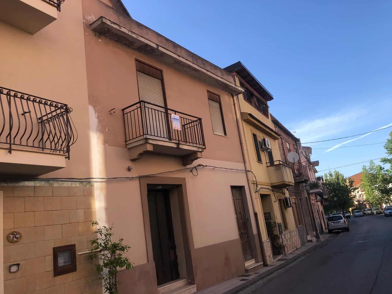 Vicolo di case residenziali con balconi e finestre, strada urbana tranquilla con veicoli parcheggiati, cielo sereno e alberi lungo il marciapiede, rappresentando un quartiere abitativo tipico italiano.