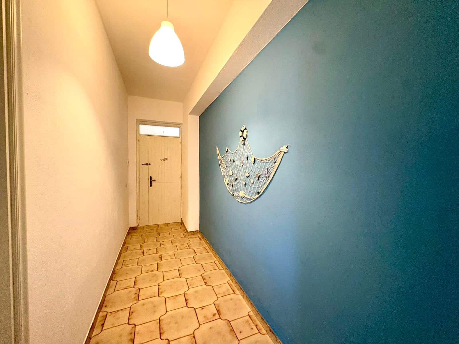 Ampia corridoio con parete blu e decorazione a forma di barca, illuminato da lampada a soffitto, con porta d'ingresso in fondo, pavimento in piastrelle beige, ambiente luminoso e accogliente.