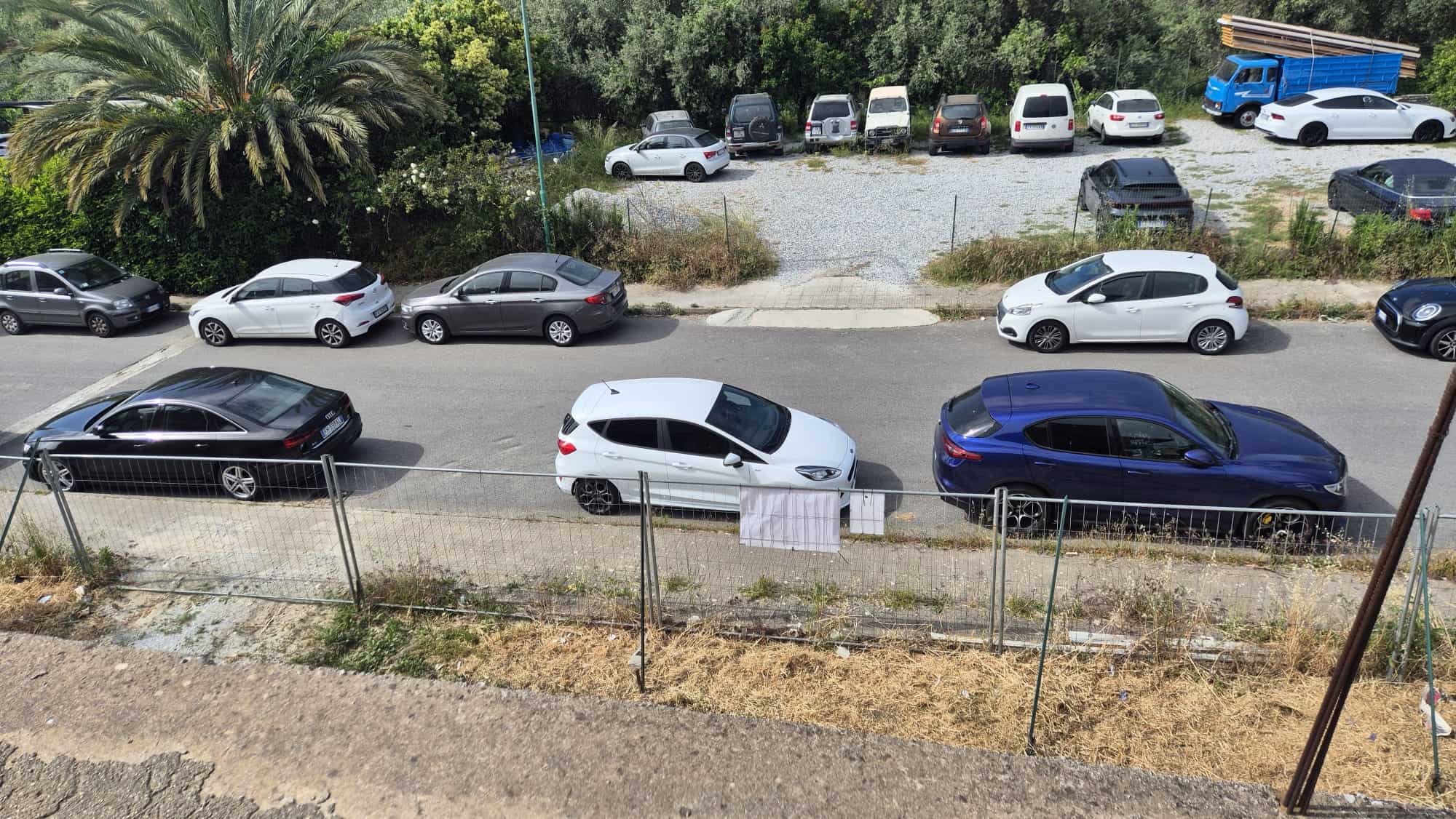 Parcheggio auto all'aperto con diverse vetture parcheggiate lungo una strada, con vegetazione e un'area di sosta in background, vista dall'alto.
