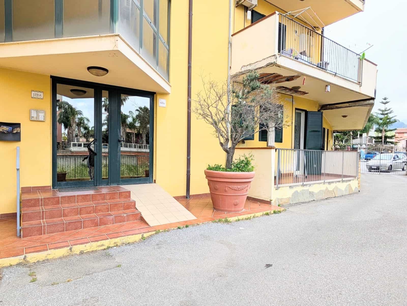 Aggiungi un'immagine di un edificio residenziale con ingresso principale, balconi e un grande vaso con pianta, in un contesto urbano soleggiato, con attenzione a dettagli come le scale e le finiture e.