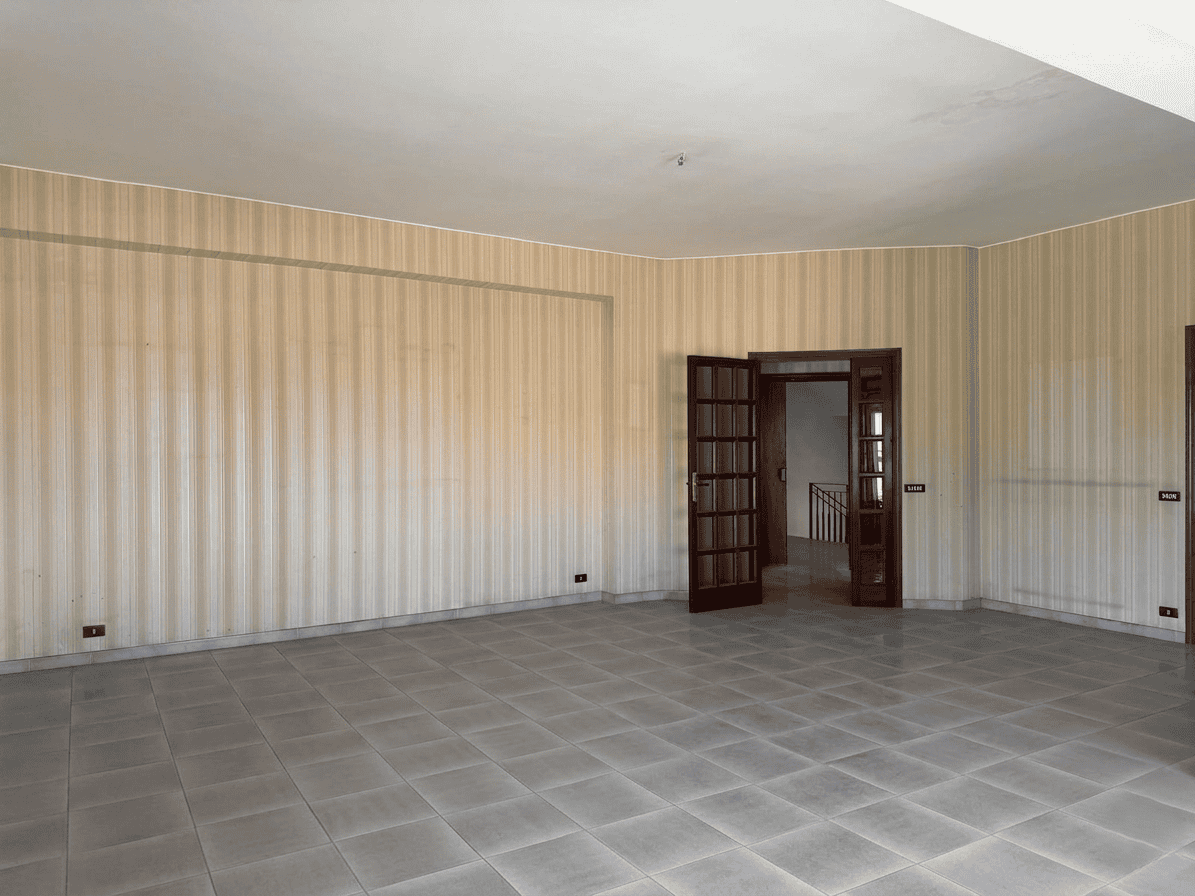 Ampio soggiorno vuoto con pareti beige e pavimento in piastrelle grigie, ingresso con porta in legno e vetri, ideale per ristrutturazione o arredamento personalizzato.
