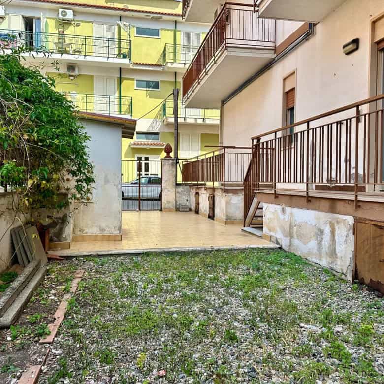 Spazio esterno di un appartamento con balcone e giardino, con edifici residenziali circostanti, pavimentazione in pietra e area verde, in un contesto urbano italiano.