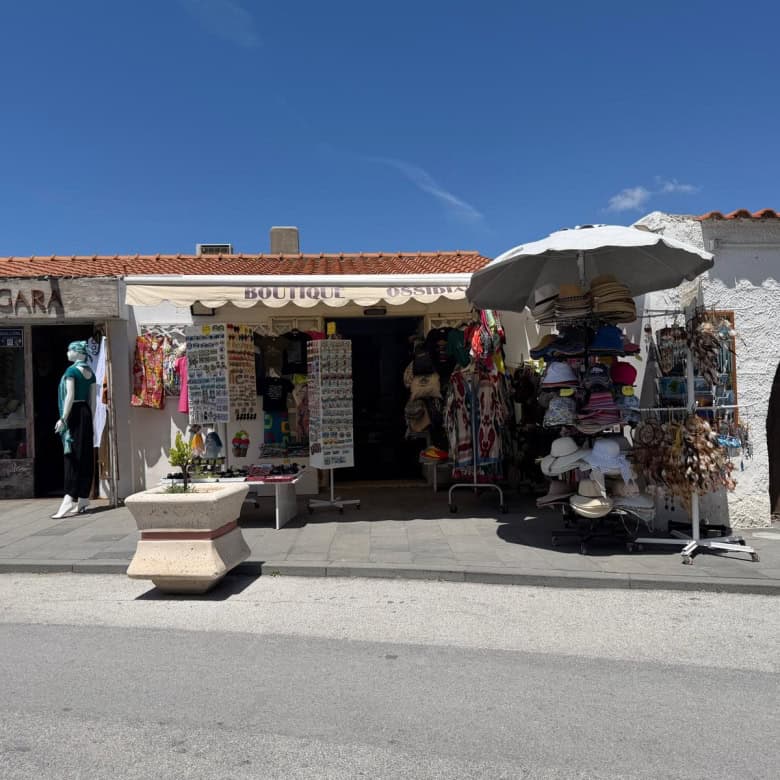 Vendita di souvenir e articoli locali in un negozio all'aperto con espositori di cappelli, abbigliamento e accessori sotto un ombrellone, in un villaggio turistico soleggiato in Italia.