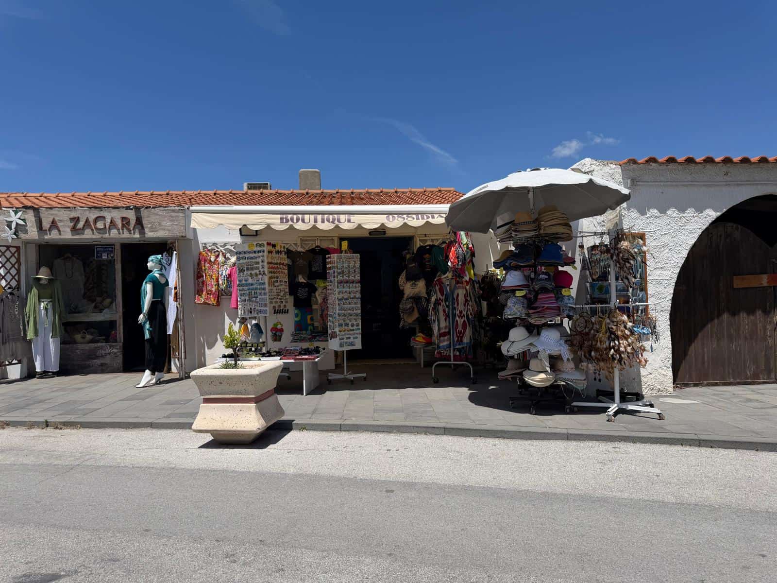 Vendita di souvenir e articoli locali in un negozio all'aperto con espositori di cappelli, abbigliamento e accessori sotto un ombrellone, in un villaggio turistico soleggiato in Italia.