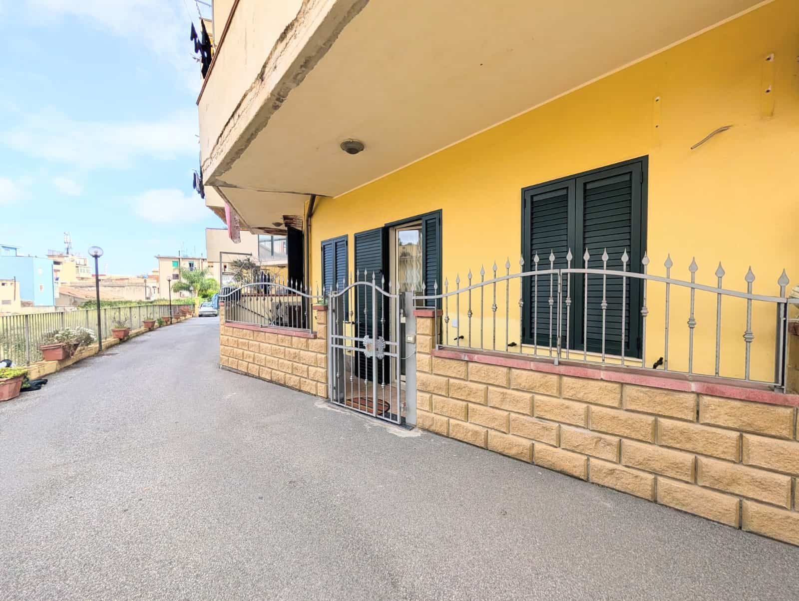 Ampia facciata di edificio residenziale con pareti gialle, finestre con persiane nere e recinzione in ferro battuto, situato in zona urbana tranquilla con strada asfaltata e spazi verdi circostanti.