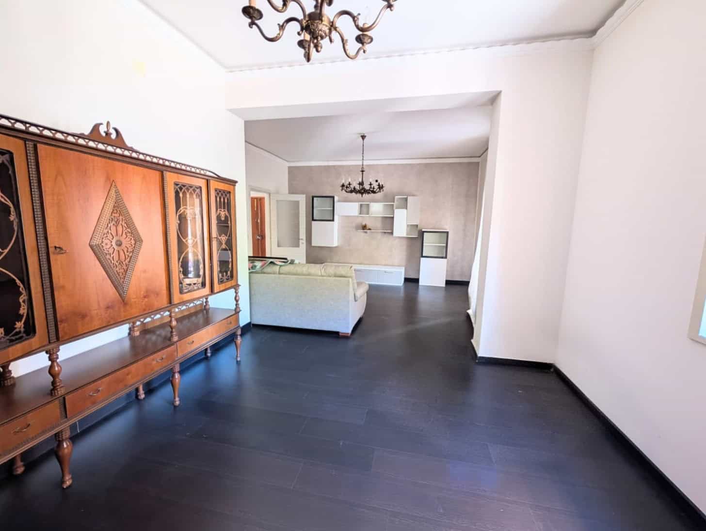 Ampio soggiorno con pavimento in legno scuro, pareti bianche e arredi classici, illuminato da lampadario centrale e luce naturale, ideale per vivere e ricevere ospiti in un ambiente elegante.