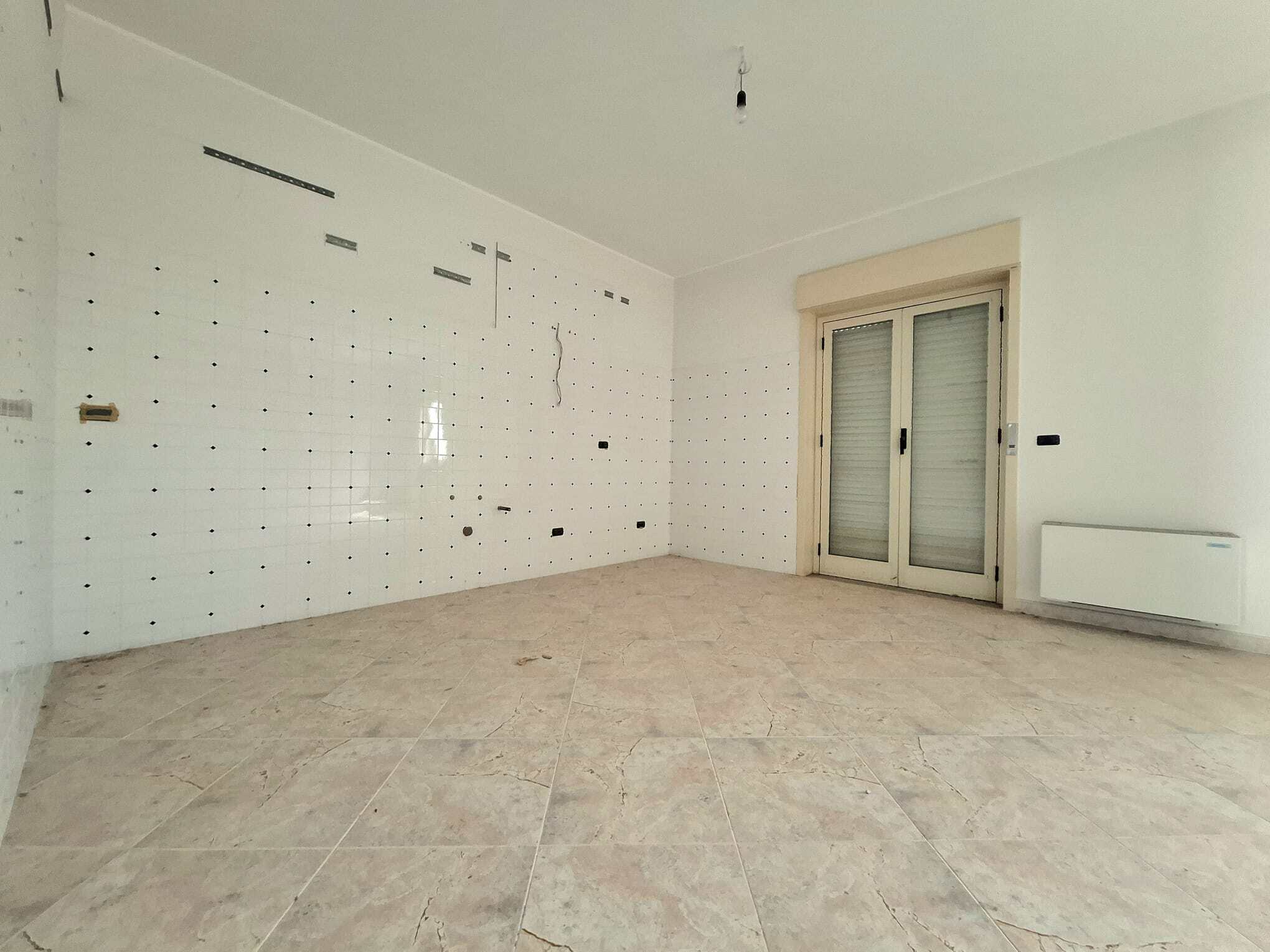 Ampia stanza vuota con pareti bianche e pavimento in marmo chiaro, grande finestra con tapparelle e radiatore, pronta per arredamento e personalizzazione.