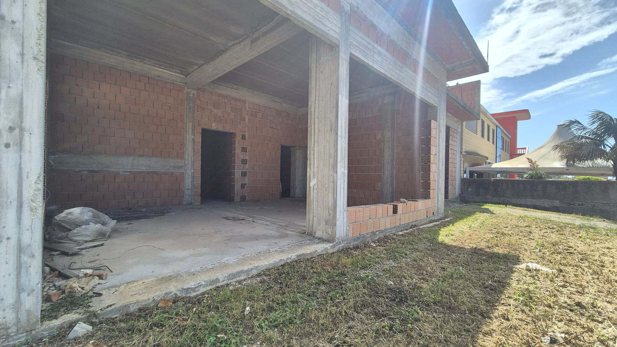 Cantiere di una casa in costruzione con struttura in cemento e mattoni, pronto per le finiture interne ed esterne, sotto un cielo sereno con alcune nuvole.