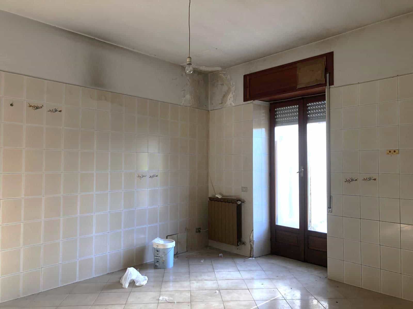 Cucina vuota in fase di ristrutturazione con pareti in piastrelle beige e porta finestra in legno, pronta per essere rinnovata.