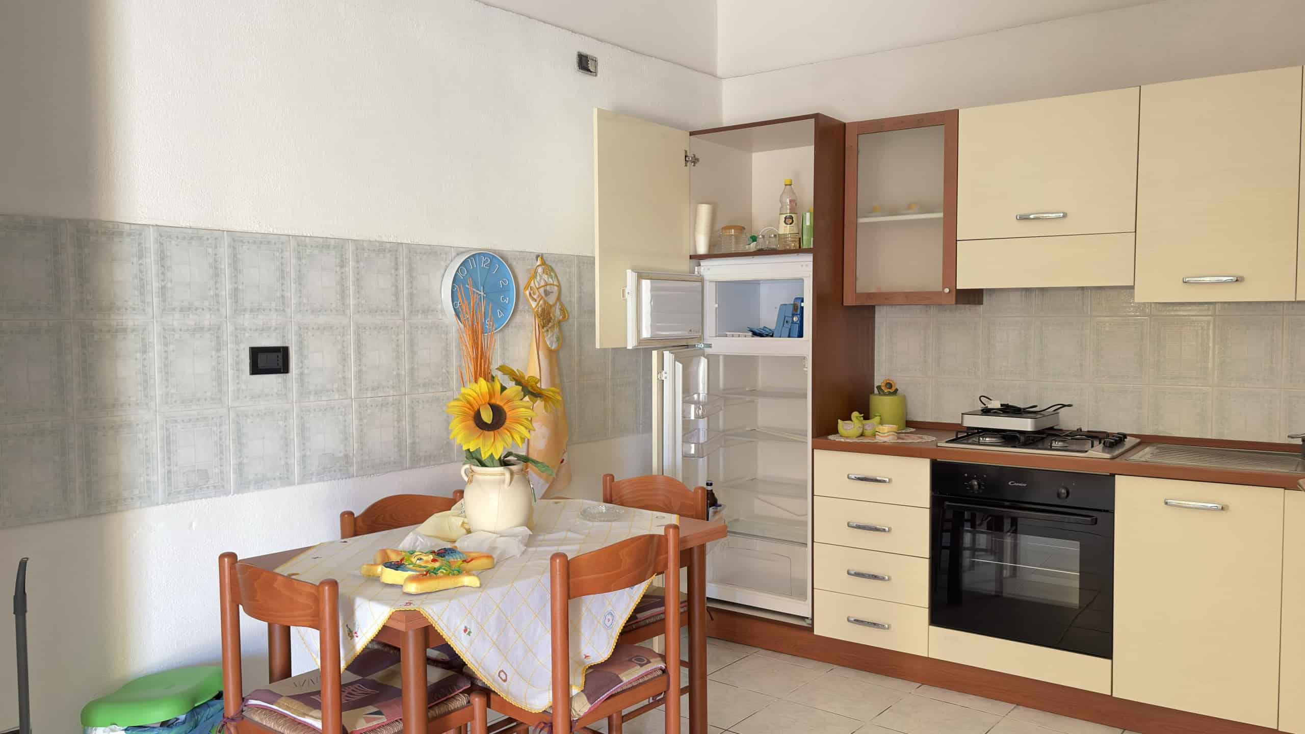 Ampia cucina moderna con elettrodomestici integrati, tavolo in legno e sedie, decorazioni floreali e frigorifero aperto, ambiente luminoso e funzionale.
