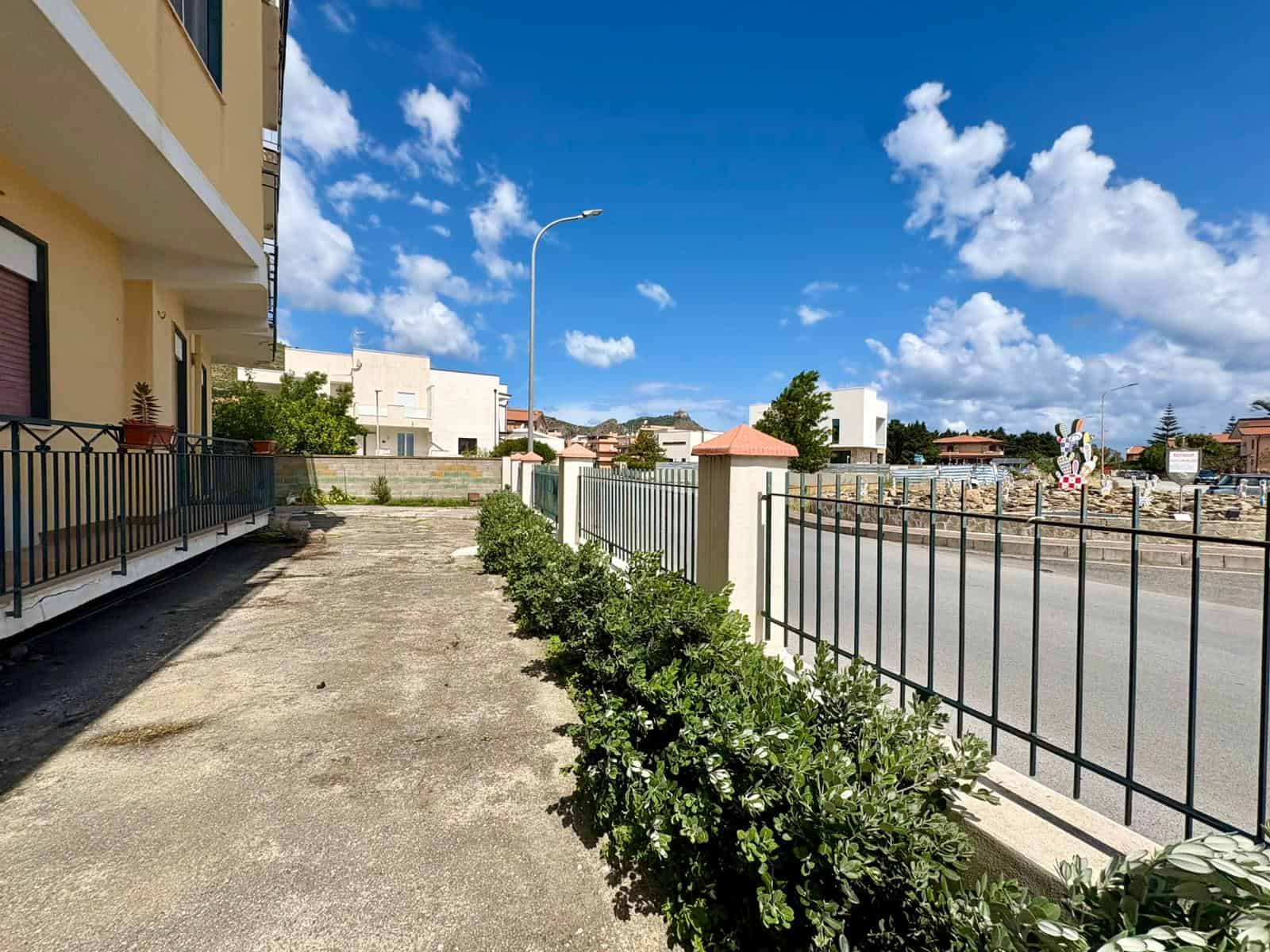 Panchina e recinzione in metallo lungo una strada urbana con edifici residenziali, cielo sereno e nuvole bianche, ambiente esterno tranquillo e ben curato, ideale per passeggiate e relax all'aperto.