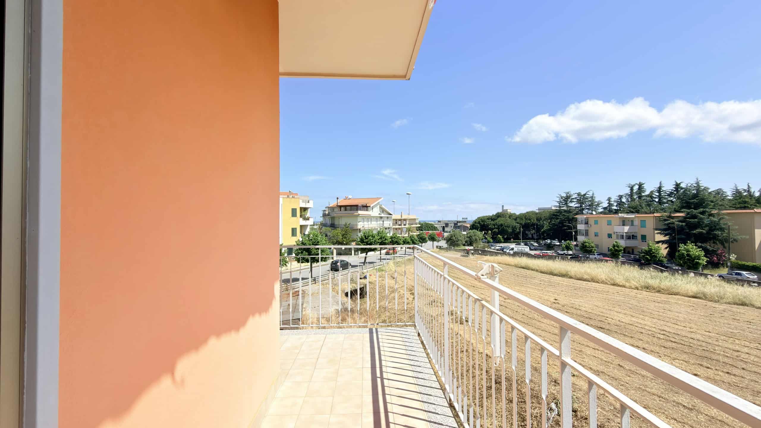 Balcone di un appartamento con vista su un'area aperta e edifici residenziali, cielo sereno e nuvole bianche, in un contesto urbano tranquillo, ideale per relax e momenti di svago all'aperto.