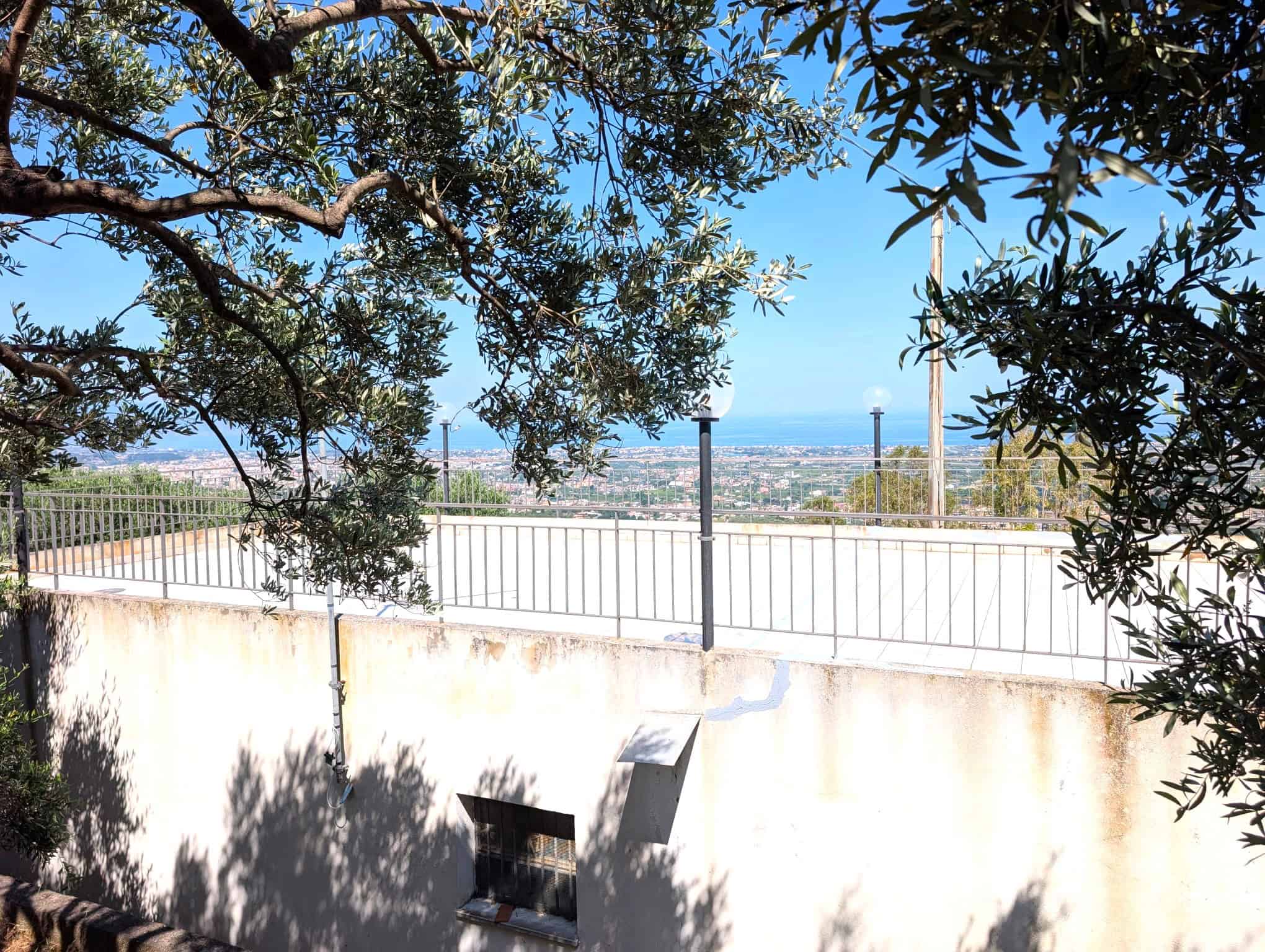 Vista panoramica dal terrazzo con vista mare, alberi e cielo azzurro, in un ambiente residenziale soleggiato e tranquillo.
