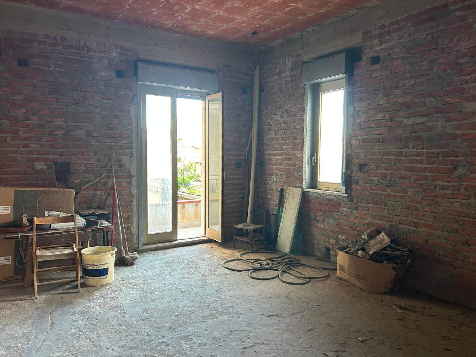 Interno di un appartamento in fase di ristrutturazione con pareti in mattoni a vista, finestre e porta finestra che permette l'accesso a un balcone, con materiali e attrezzi da lavoro presenti.