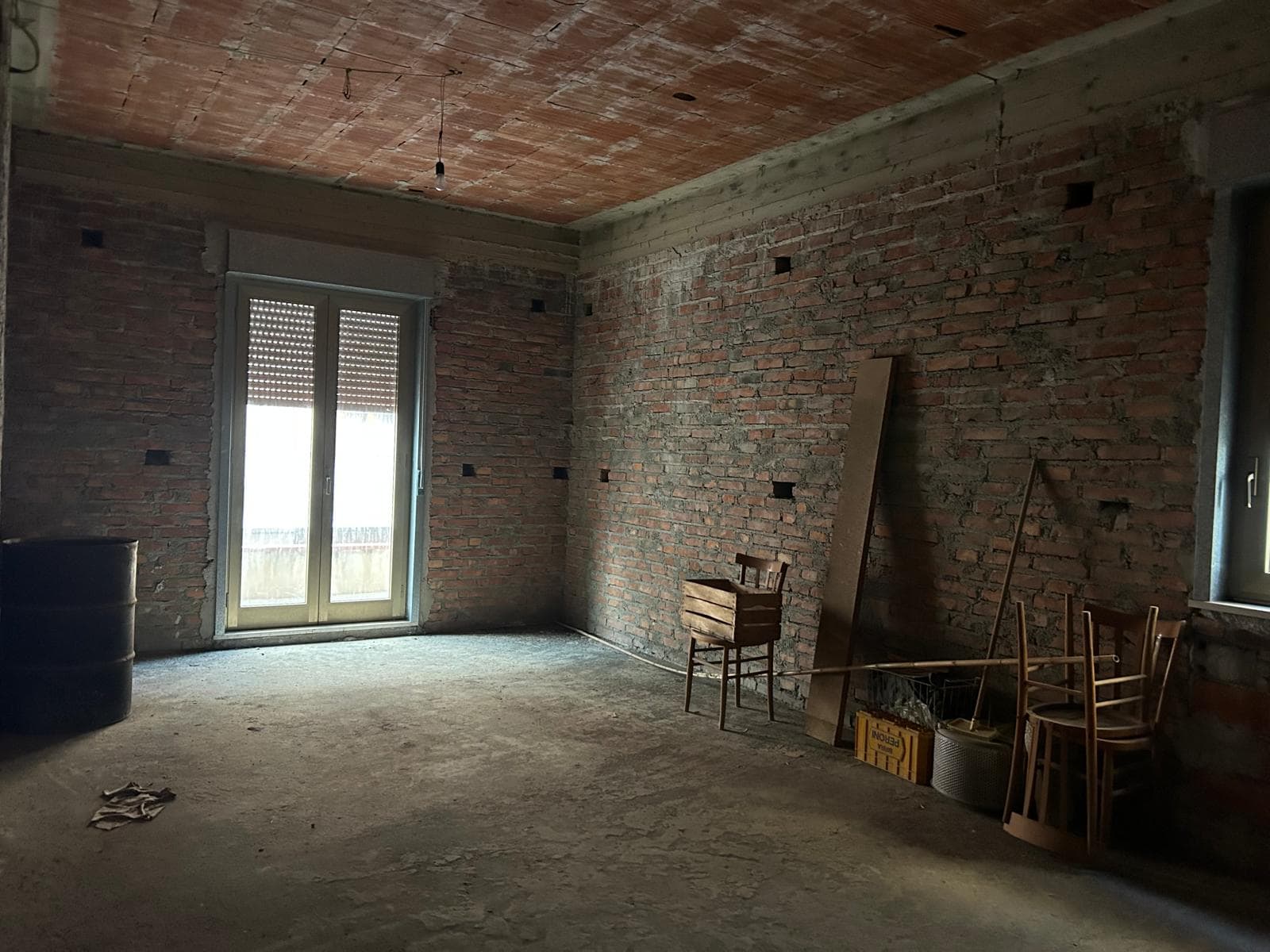 Interno di un appartamento in fase di ristrutturazione con pareti in mattoni a vista e grandi finestre che lasciano entrare molta luce naturale.