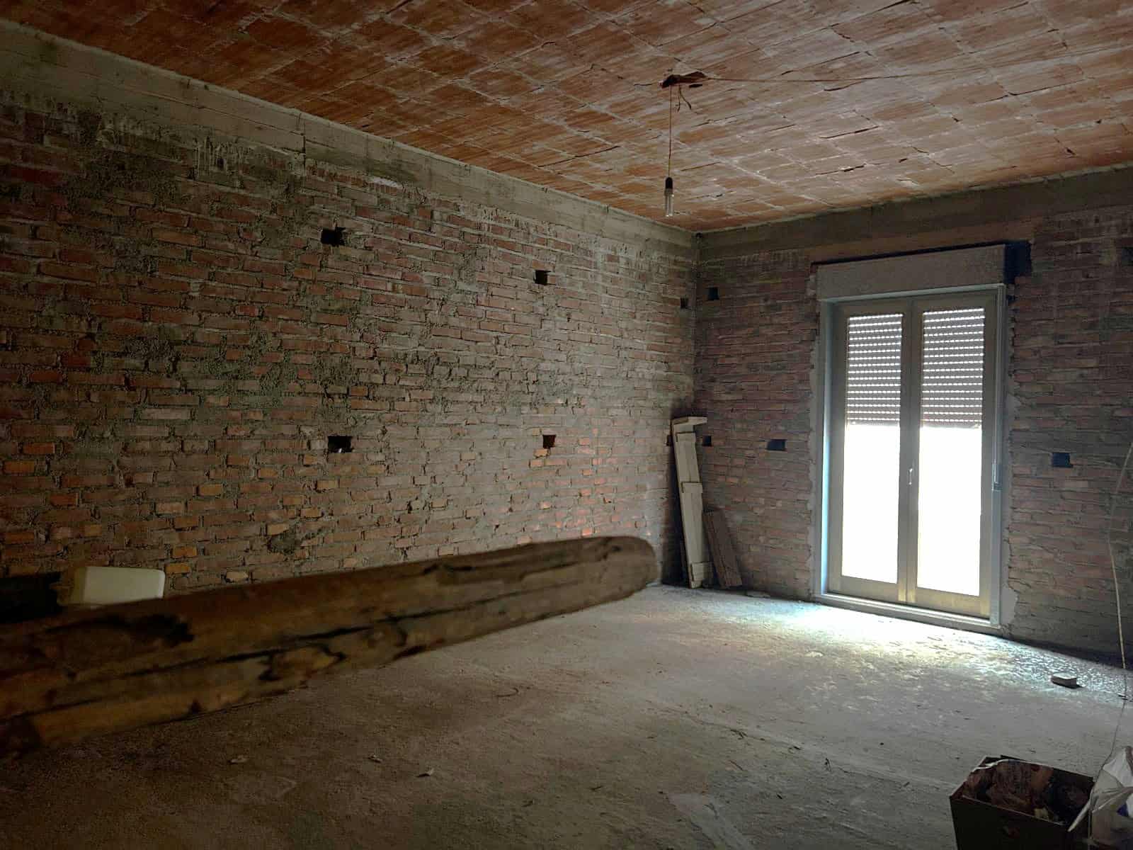 Interno di un appartamento in fase di ristrutturazione con pareti in mattoni a vista e grande finestra con tapparelle, pronto per essere arredato.