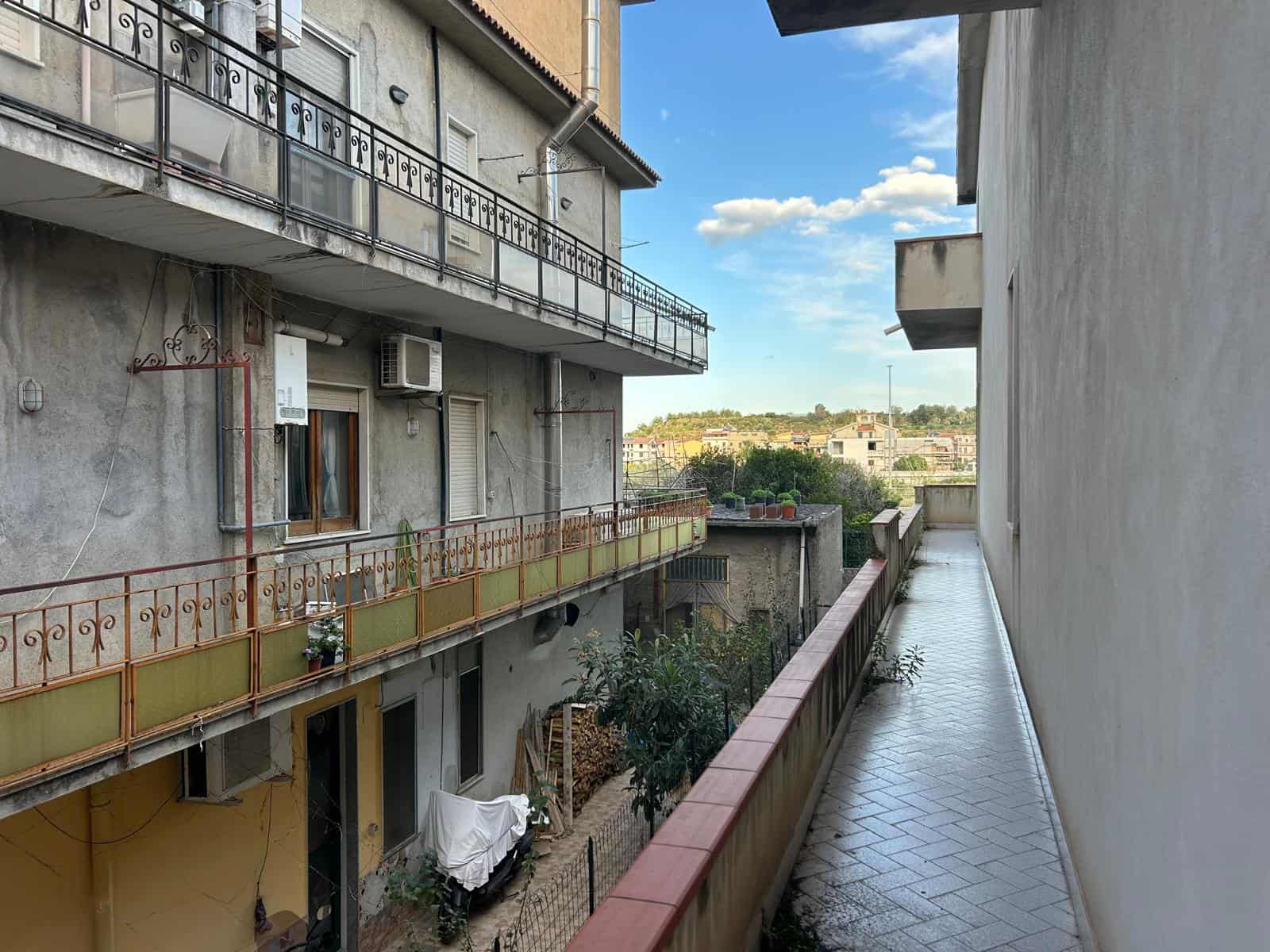 Un balcone di un edificio residenziale con vista sul cielo azzurro e le nuvole, con piante e oggetti vari, in un contesto urbano.