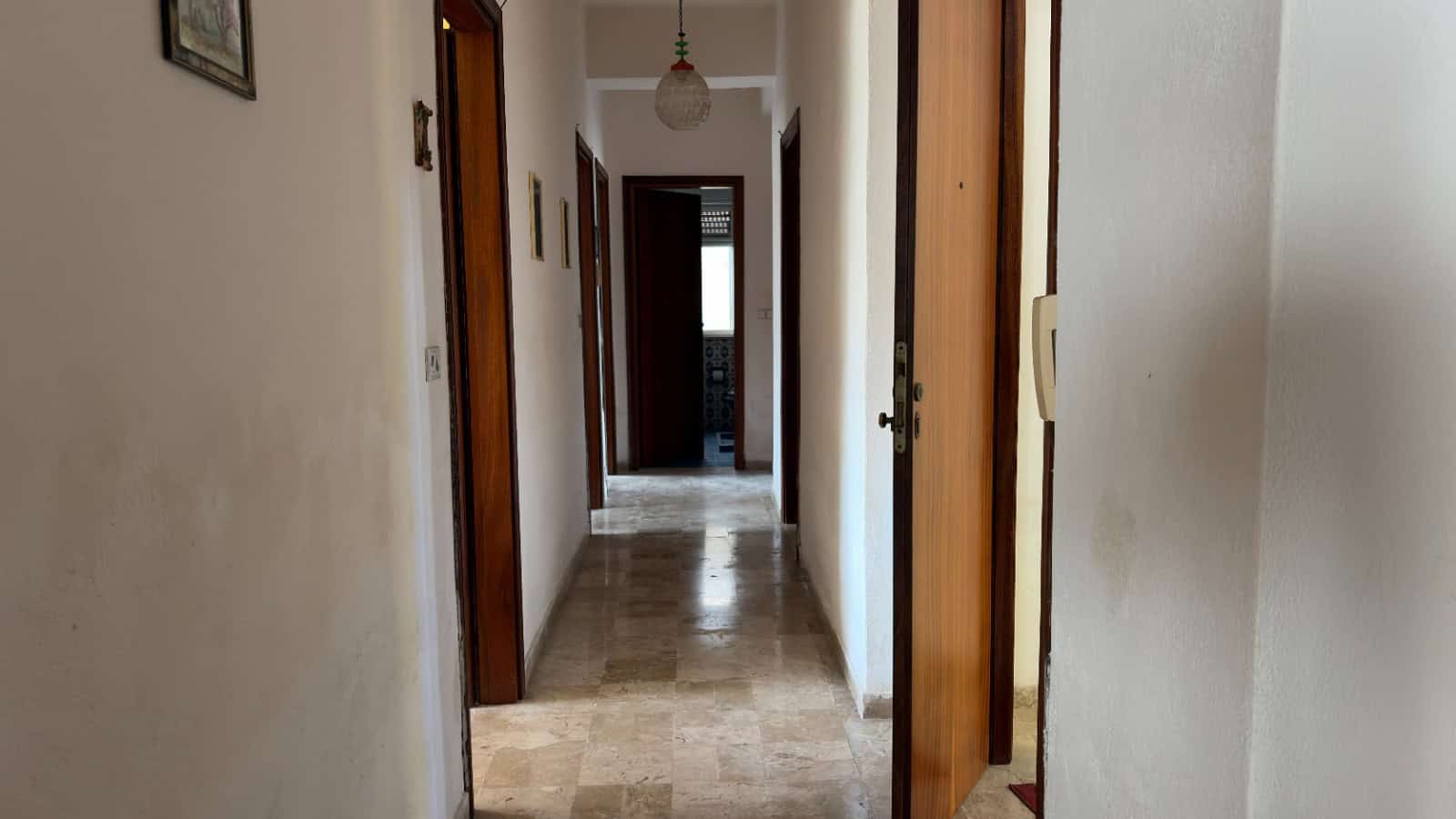 Ampia vista di un corridoio interno di una casa con pareti bianche e pavimento in marmo beige, con porte in legno scuro e decorazioni alle pareti.
