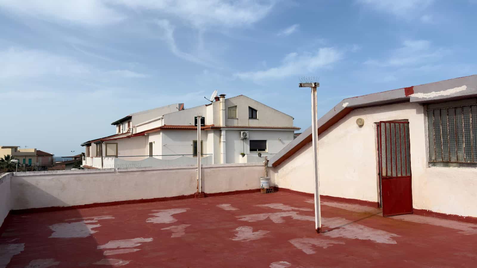 Balcone panoramico con vista sul cielo azzurro e case circostanti, superficie in cemento rosso, ringhiera in metallo e porta in legno, ambiente esterno tranquillo e luminoso.
