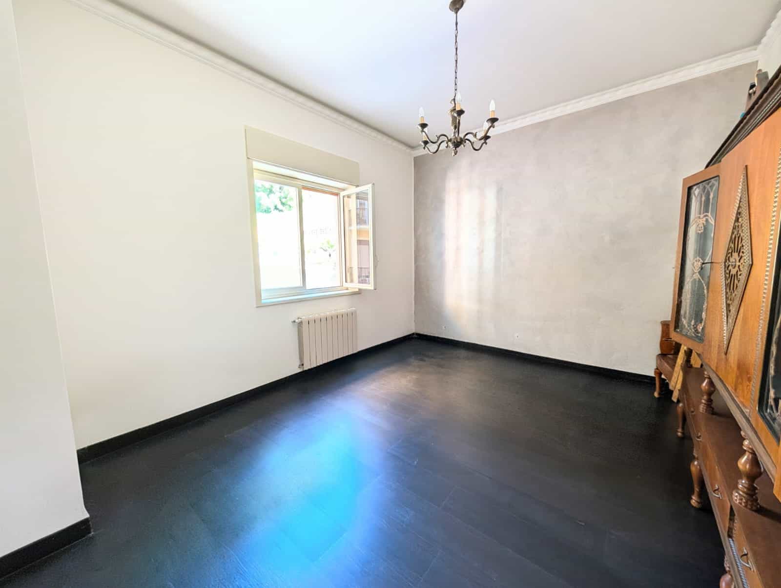 Ampio soggiorno luminoso con pareti bianche, grande finestra e pavimento in legno scuro, ideale per arredamento moderno e accogliente.