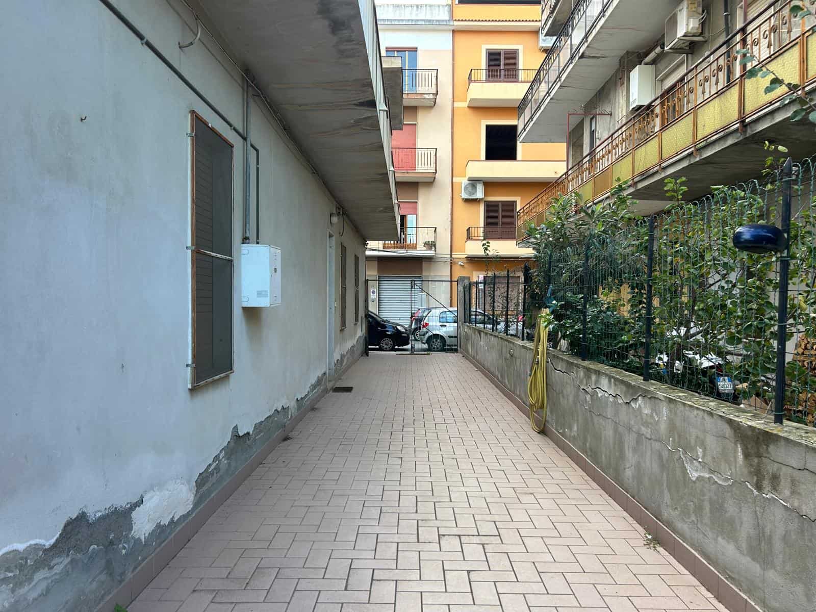 Corridoio esterno tra edifici residenziali con pavimentazione in mattonelle e piante lungo il lato destro, in zona urbana.