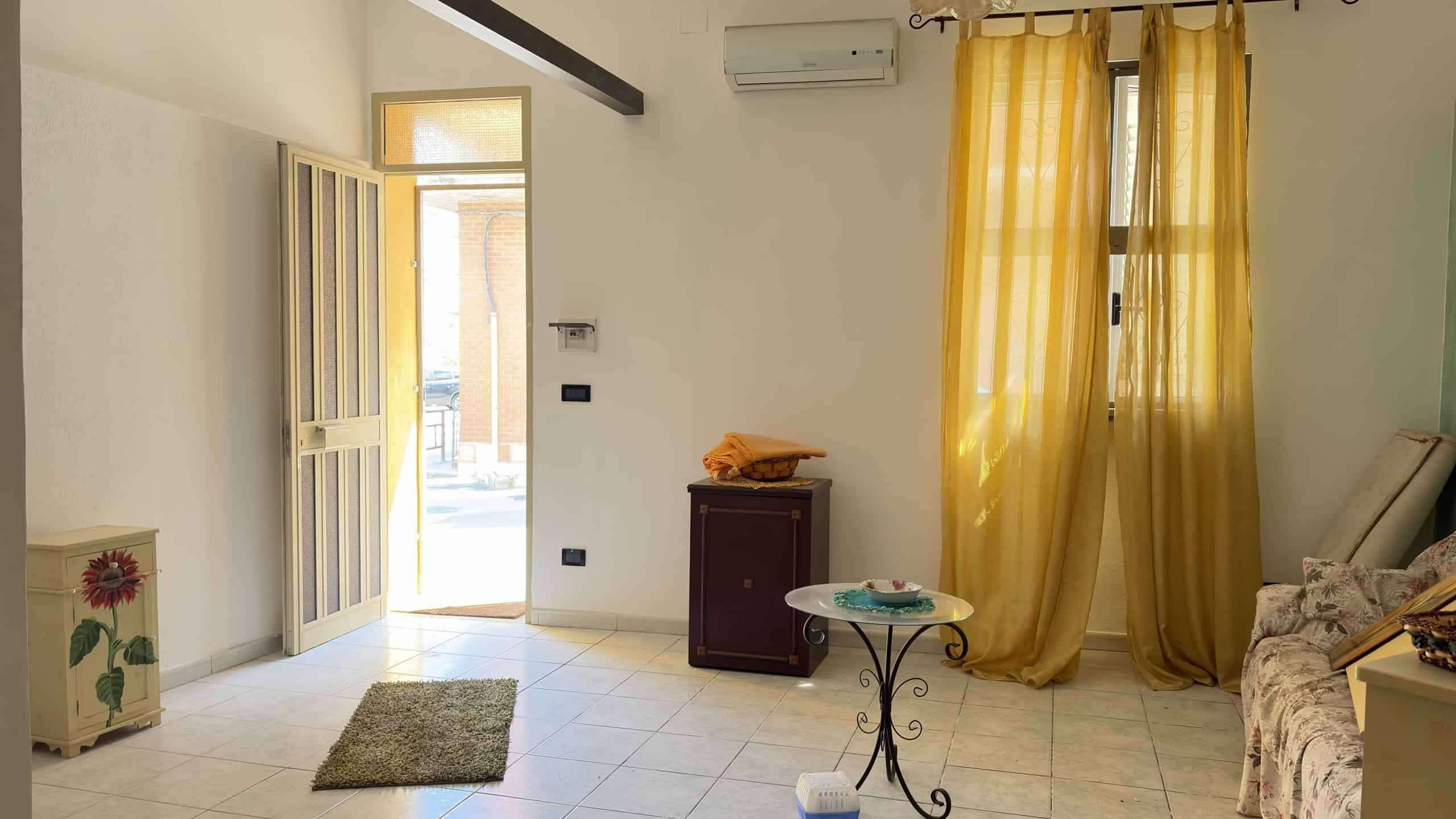 Porte d'ingresso aperta in una casa luminosa con tende gialle, arredamento semplice e pavimento in piastrelle bianche, creando un ambiente accogliente e luminoso.