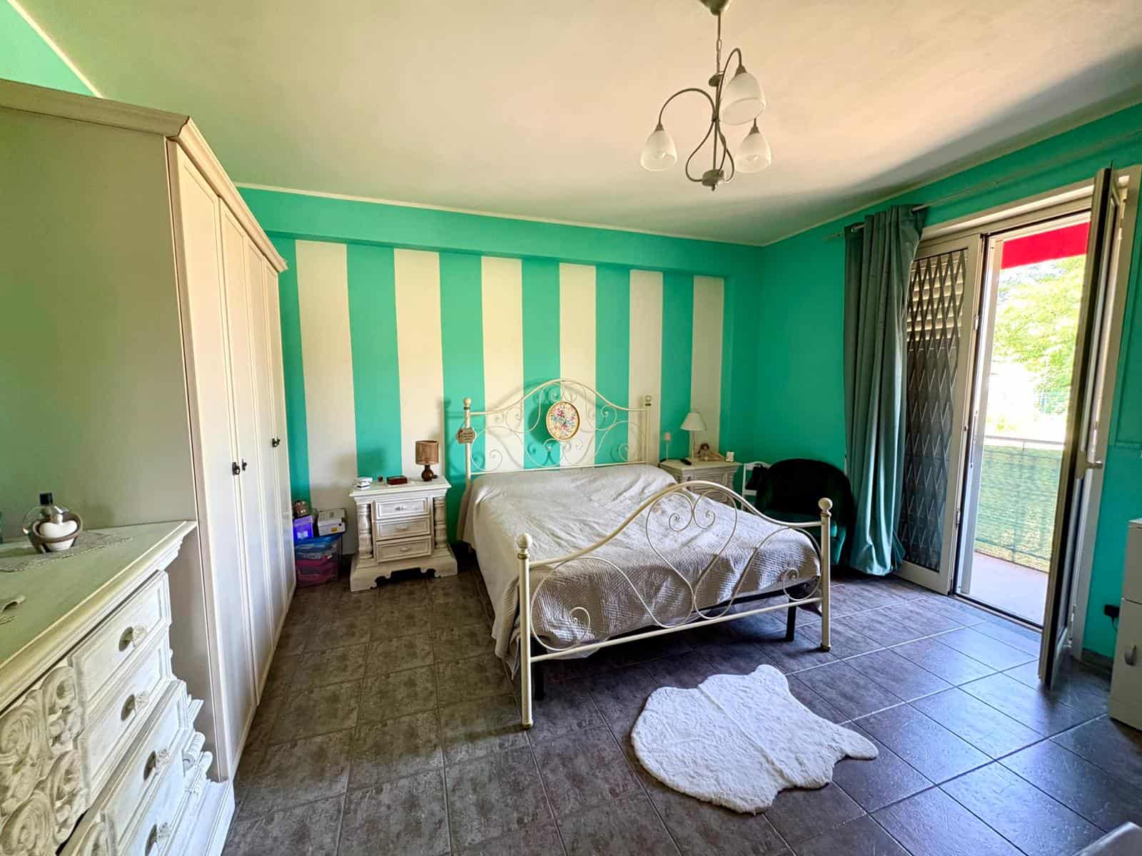 Letto matrimoniale in una camera da letto luminosa con pareti a righe verde acqua e bianco, armadio bianco e accessori decorativi.