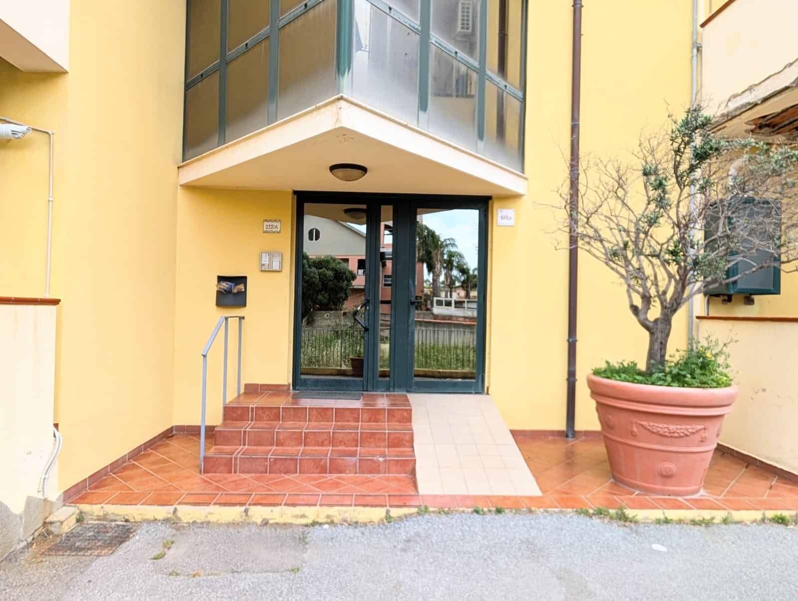 Ampio ingresso condominiale con porta in vetro e scala accessibile, facciata gialla con piante ornamentali, ambiente luminoso e accogliente, ideale per appartamenti moderni e confortevoli.