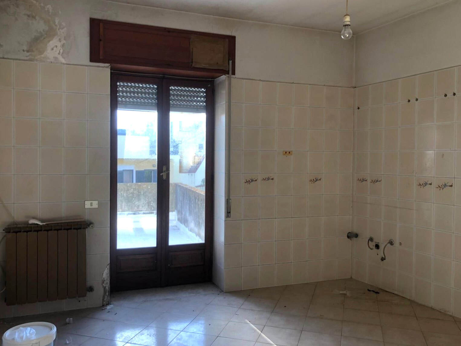 Cucina vuota in fase di ristrutturazione con pareti in piastrelle beige e porta finestra che dà su un balcone, pronto per essere arredato.