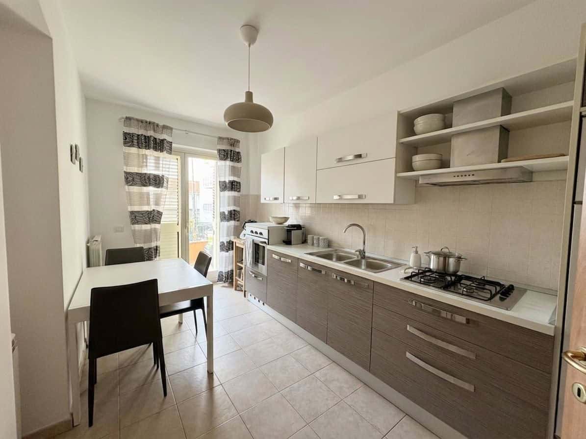 Cucina moderna e luminosa in appartamento NextCasa con mobili bianchi e grigi, piano cottura a gas e zona pranzo con finestra con tende a righe.