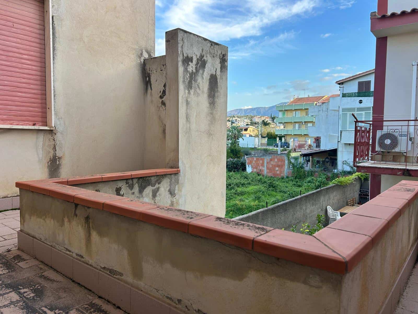 Balcone di un appartamento con vista su edifici residenziali e spazi verdi, in un contesto urbano mediterraneo, con cielo sereno e clima soleggiato, ideale per relax e momenti all'aperto.