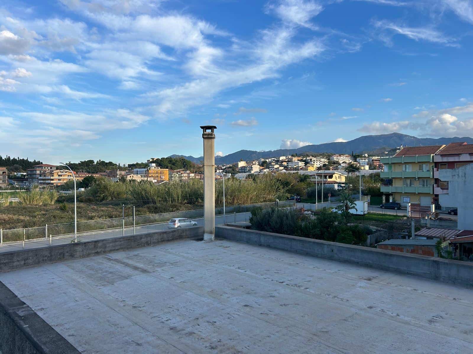 Il cielo azzurro con nuvole bianche sopra un'area urbana con edifici e montagne sullo sfondo, vista da un terrazzo o tetto di un edificio.