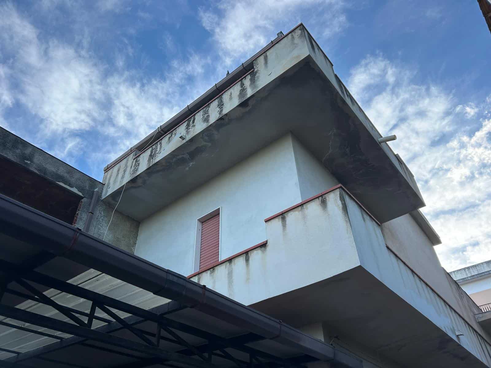Balcone di un edificio residenziale con segni di usura e ruggine, vista dall'angolazione verso il cielo con nuvole bianche e azzurre, evidenziando l'aspetto esterno e lo stato di manutenzione della st.