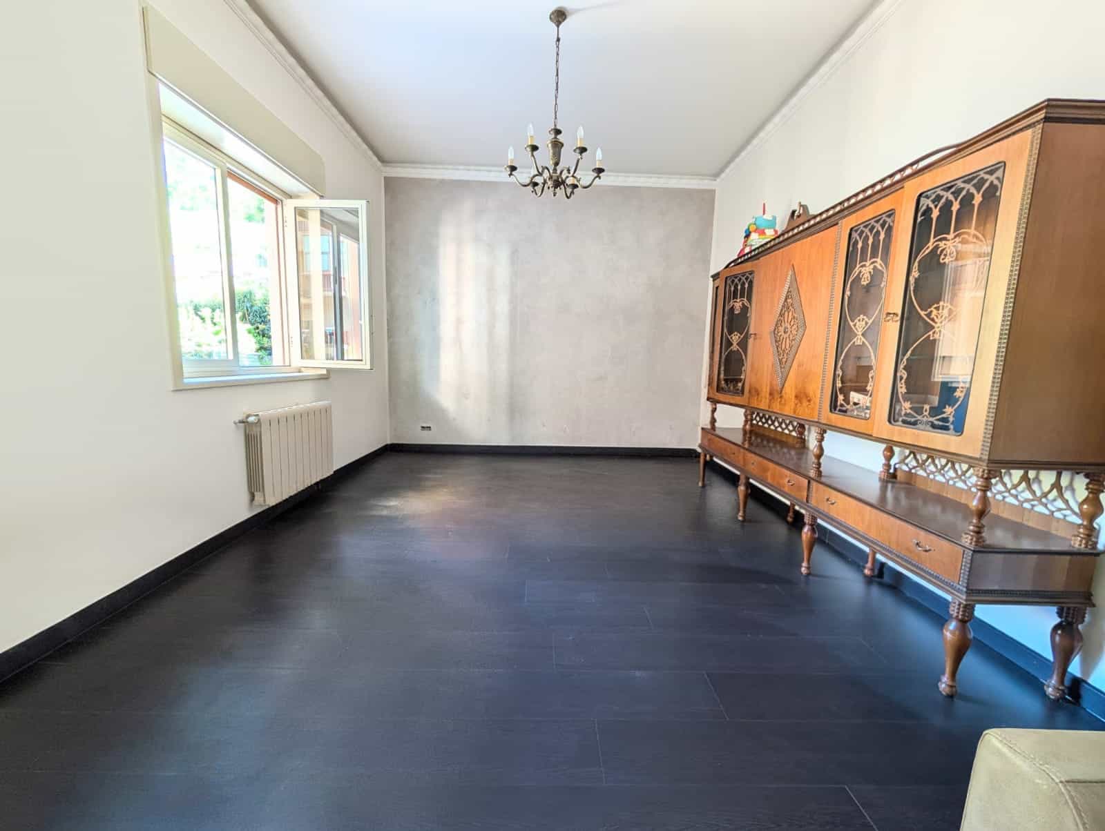 Sala spaziosa con pareti bianche, grande finestra e mobile antico in legno, illuminata da lampadario centrale, ideale per soggiorno o sala da pranzo.
