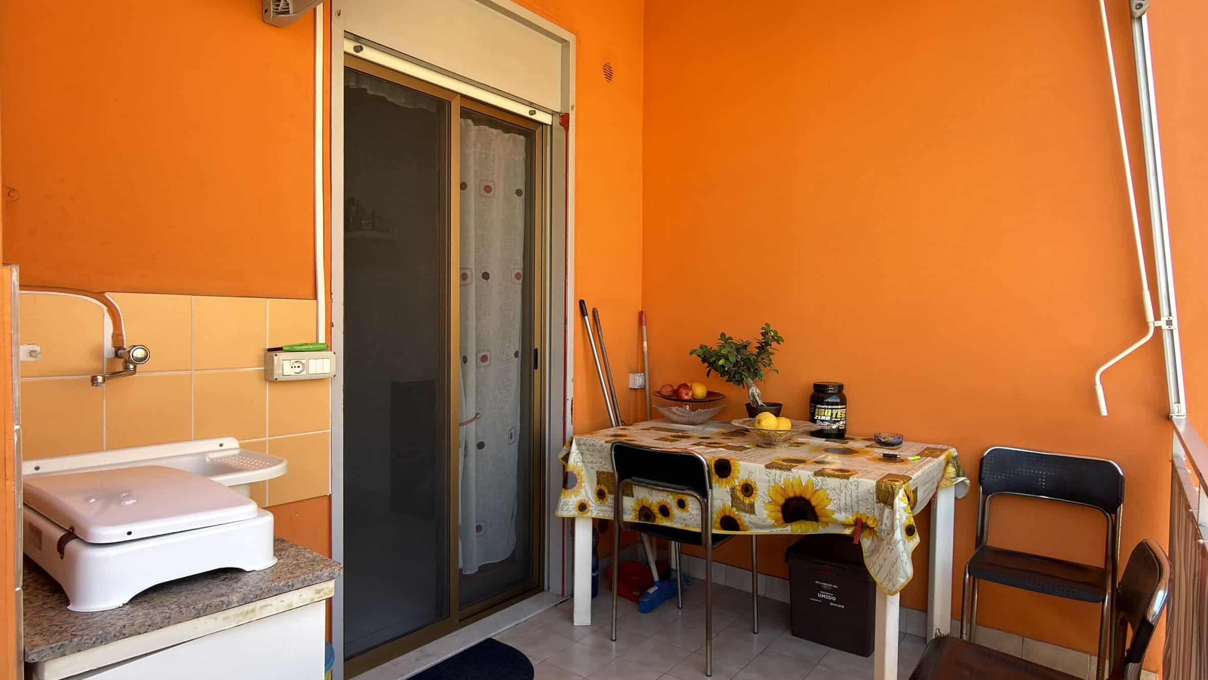Ampio balcone con porta scorrevole, tavolo con tovaglia a fiori, sedie, piante e oggetti decorativi, in un ambiente luminoso e accogliente con pareti arancioni.
