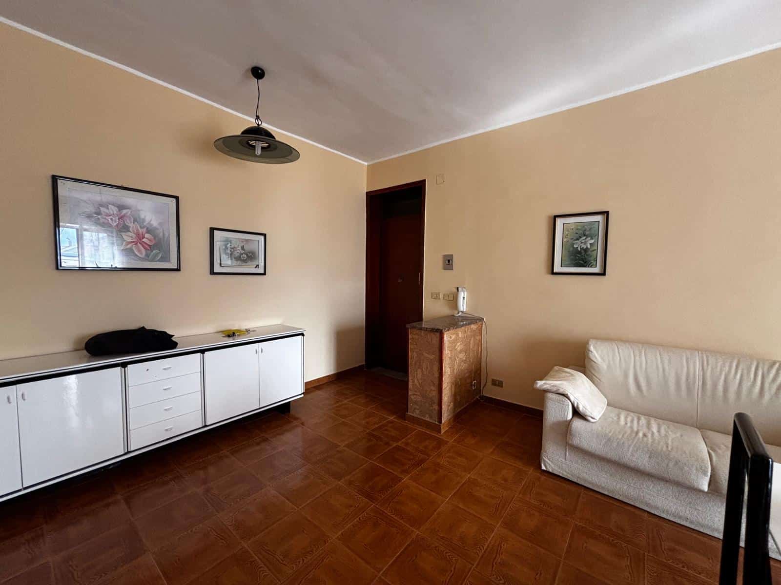 Interno di soggiorno con pareti beige, divano bianco, mobili bianchi e decorazioni floreali alle pareti, ambiente luminoso e accogliente.