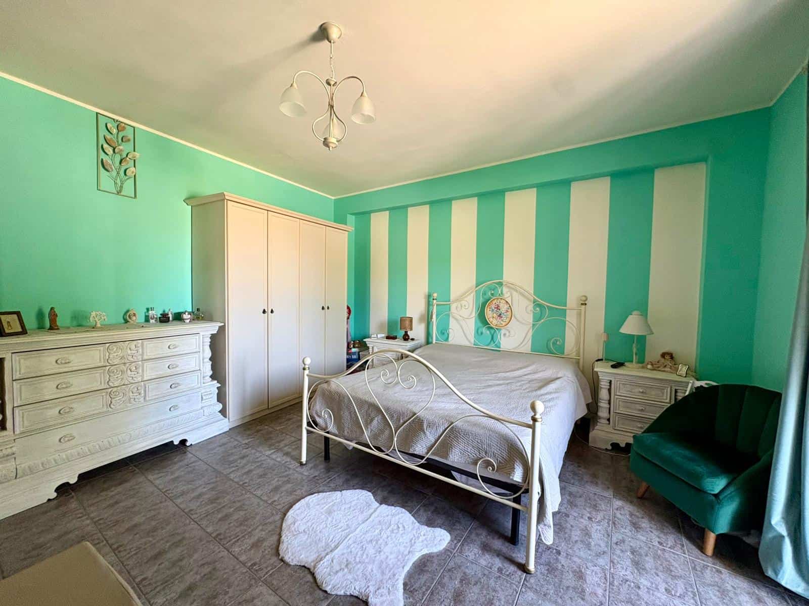 Camera da letto luminosa con pareti a righe verde acqua e bianco, arredi eleganti in stile vintage, armadio bianco, letto con testiera in ferro battuto, e poltrona verde scuro, ambiente accogliente e.