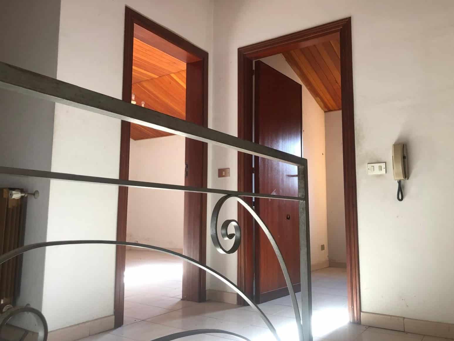 Vista dell'ingresso di una casa con pareti bianche, porte in legno scuro e soffitto in legno, con ringhiera in metallo decorativa, ambiente luminoso e accogliente.