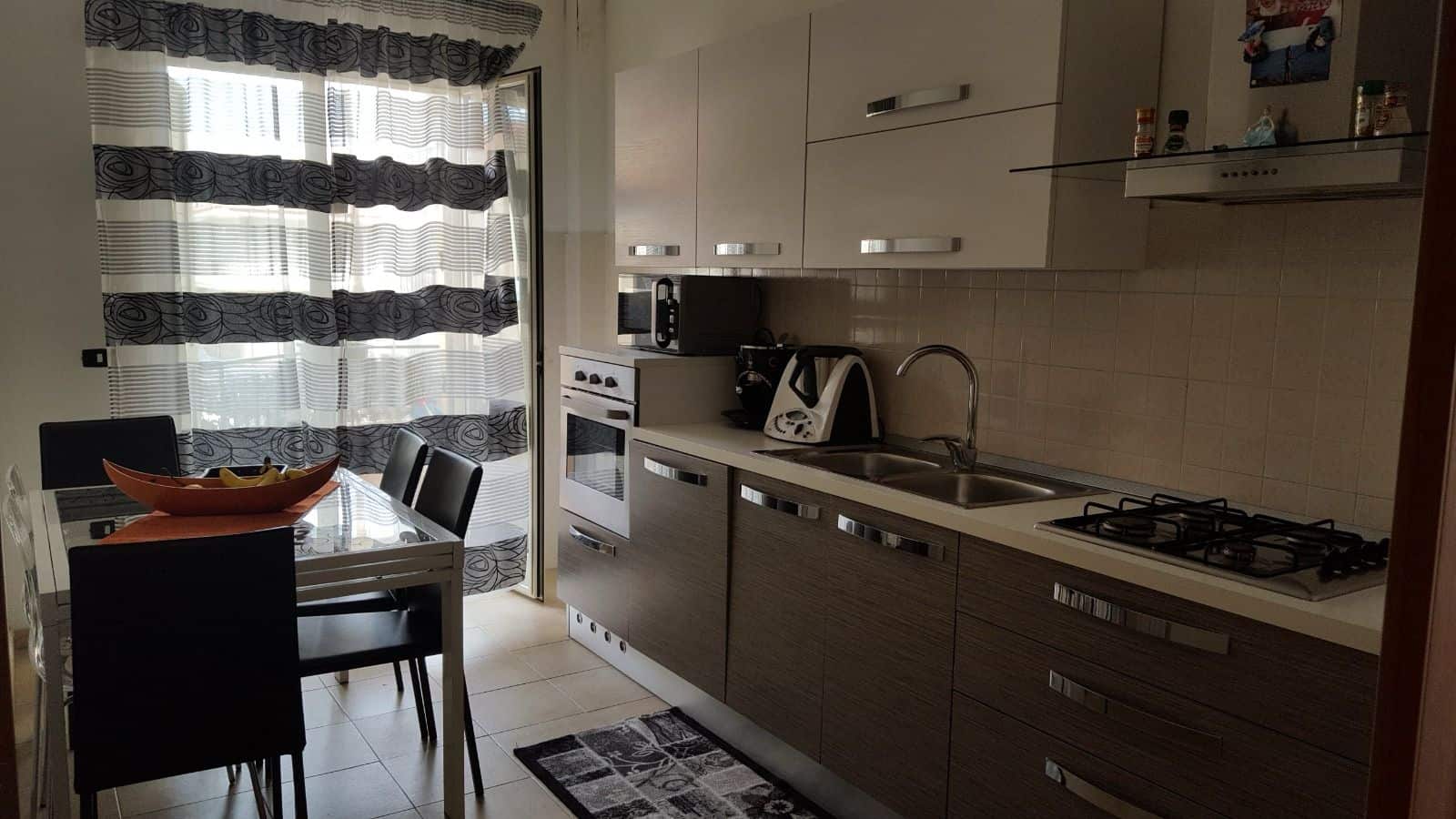 Cucina moderna con mobili beige e elettrodomestici integrati, illuminata da luce naturale attraverso una finestra con tende a righe.