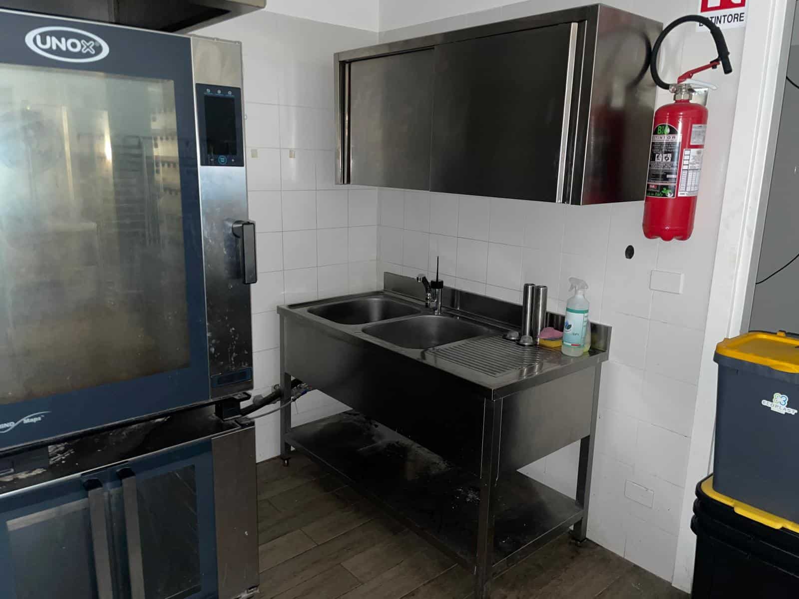 Piatto di cucina professionale con lavello in acciaio inox, forno industriale e armadietti di stoccaggio in ambiente di ristorante o cucina commerciale.
