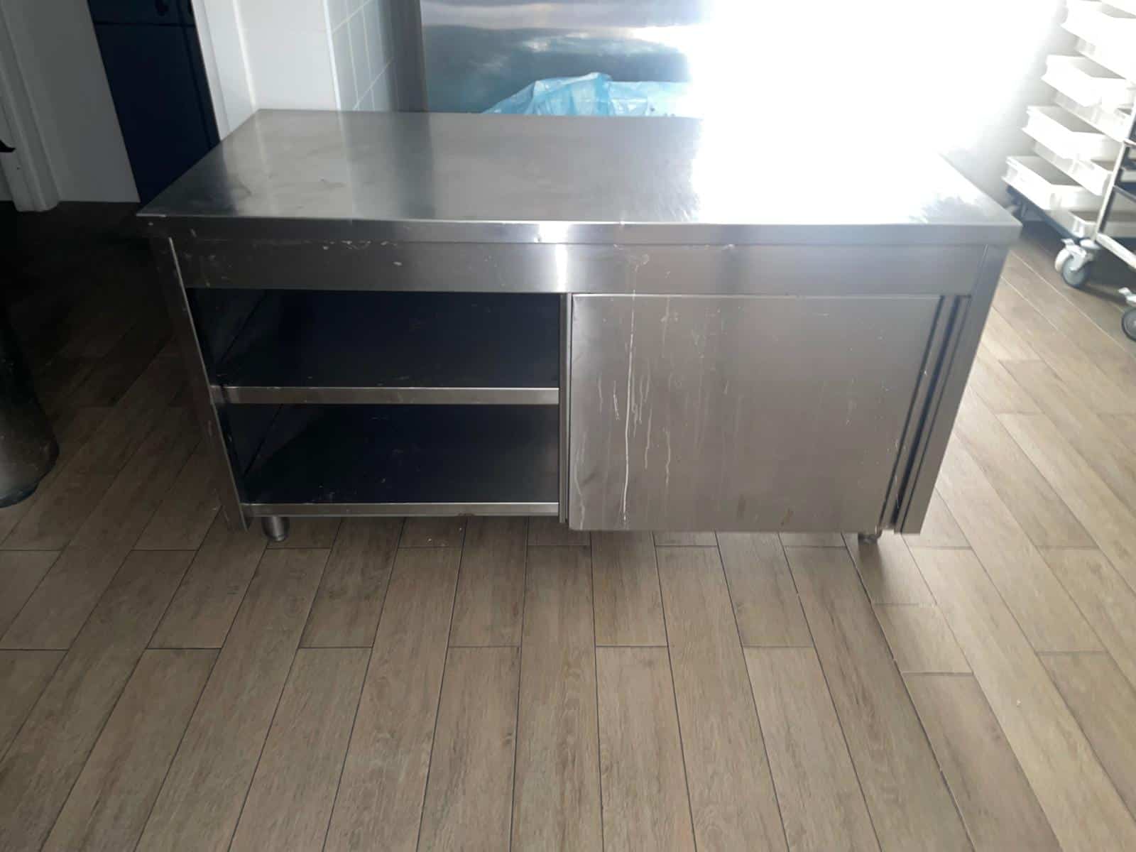Una scrivania in acciaio inossidabile in un ambiente professionale, con superficie liscia e spazio di archiviazione aperto, ideale per cucine industriali o laboratori di cucina.