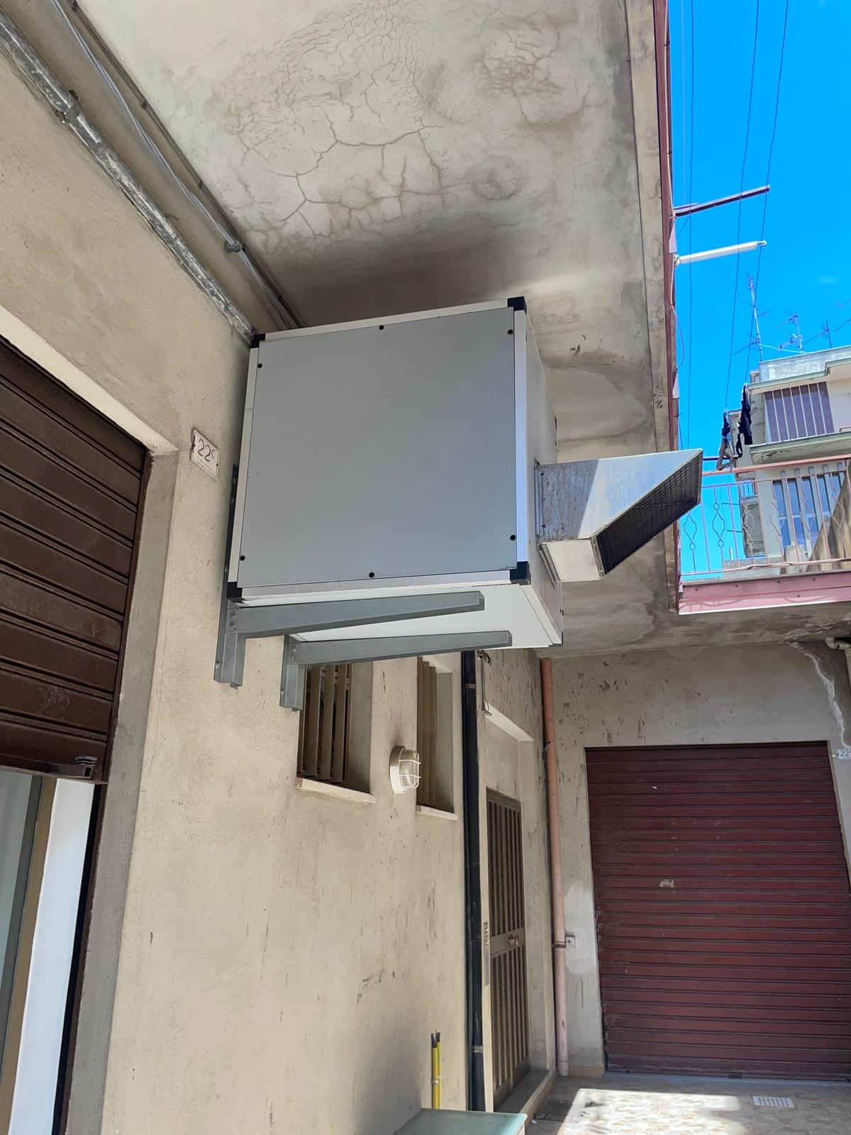 Sistema di climatizzazione esterno installato su parete di un edificio, con tubazioni e unità di condizionamento visibili, in un contesto urbano con balcone e serrande.