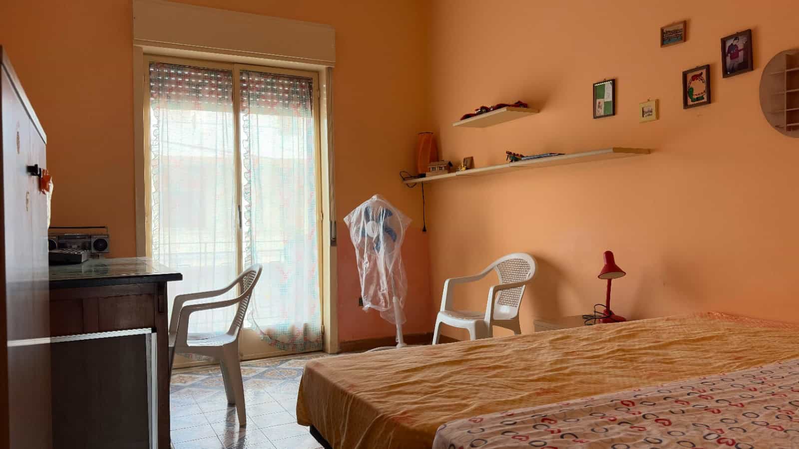 Una camera da letto luminosa con pareti color pesca, letto matrimoniale, sedie e scaffali con decorazioni, finestra con tende trasparenti che lascia entrare la luce naturale.