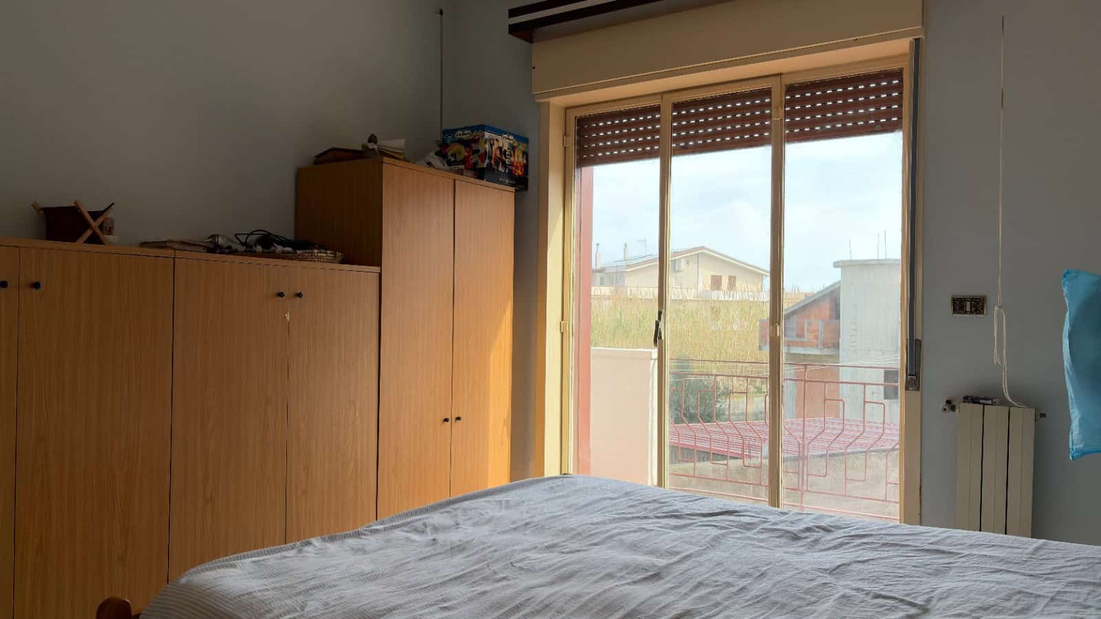 Ampia camera da letto con armadio in legno e grande finestra che permette l'ingresso di luce naturale, vista su edifici e balcone, ideale per immobili residenziali di NextCasa.