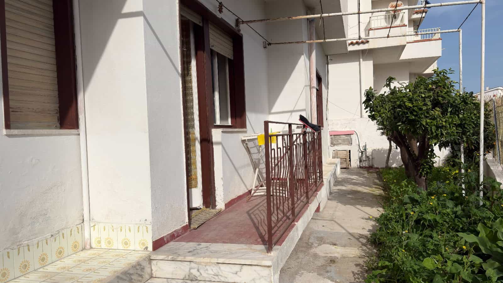 Balcone di un appartamento con ringhiera in metallo, accesso tramite porta finestra, con piante e alberi sullo sfondo, in un contesto residenziale soleggiato.
