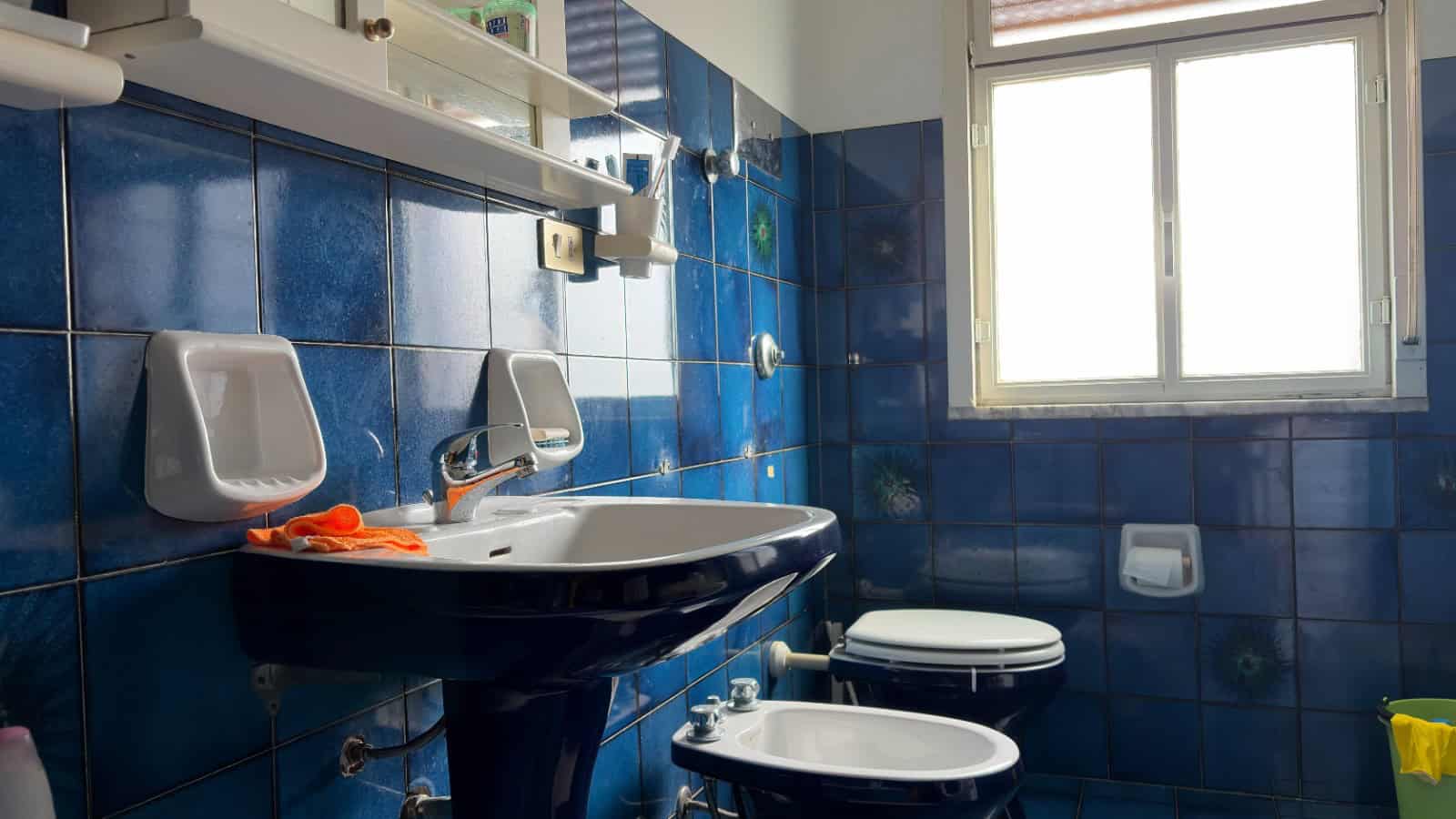 Lavandino e servizi igienici in bagno con piastrelle blu, finestra luminosa e accessori essenziali, ideale per soluzioni abitative moderne e funzionali.