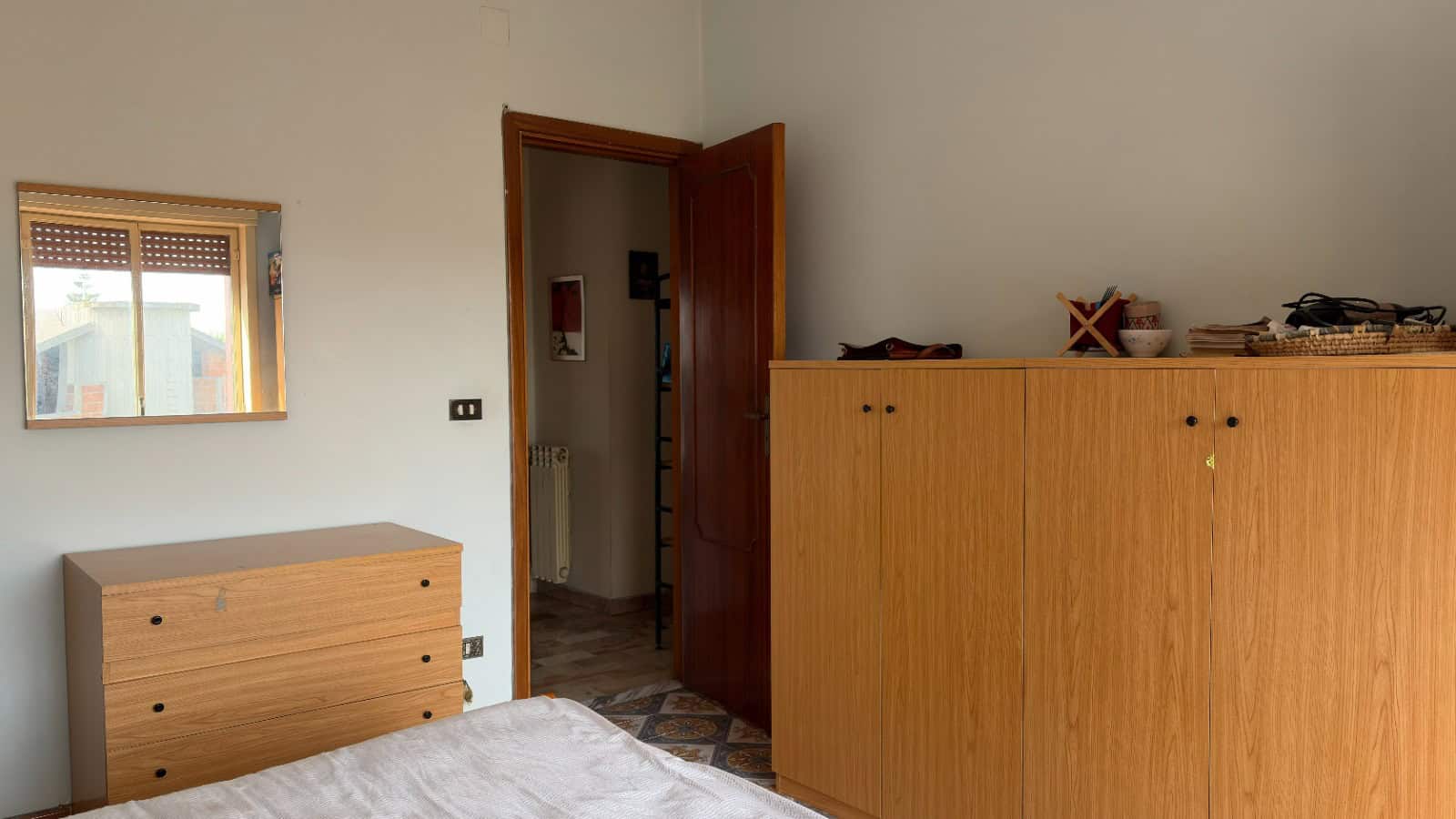 Armadio e cassettiera in legno in una stanza da letto con pareti bianche e pavimento in piastrelle.
